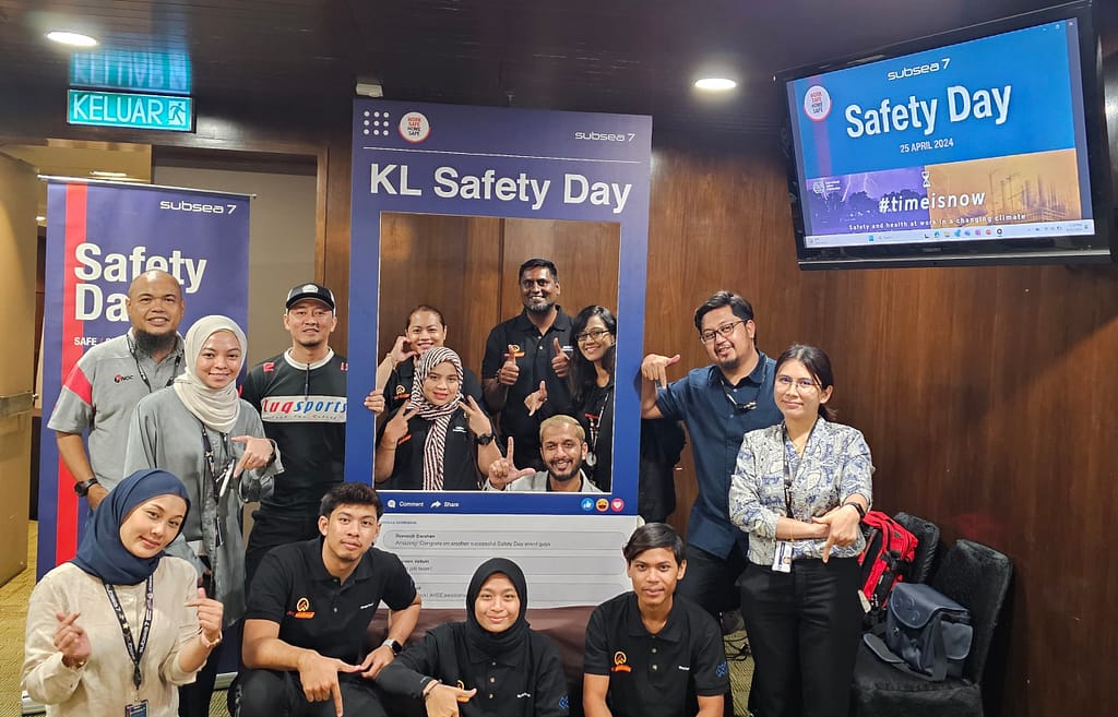 Group of diverse participants posing for KL Safety Day event, featuring safety banners and a digital display, emphasizing workplace safety and health.
