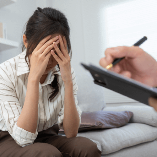 Woman with hands on her face expressing distress during a psychological assessment session, with a psychologist taking notes in the background.