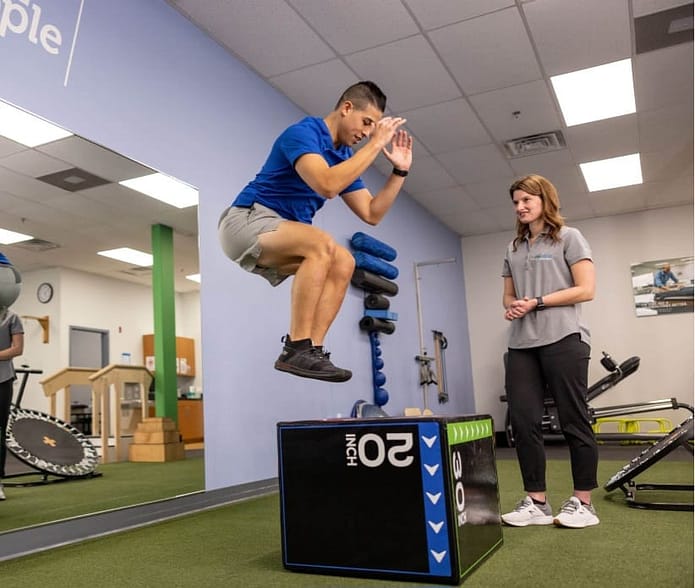 Athlete performing jump exercise on a 20-inch box during sports rehabilitation session, with therapist observing in fitness center setting.