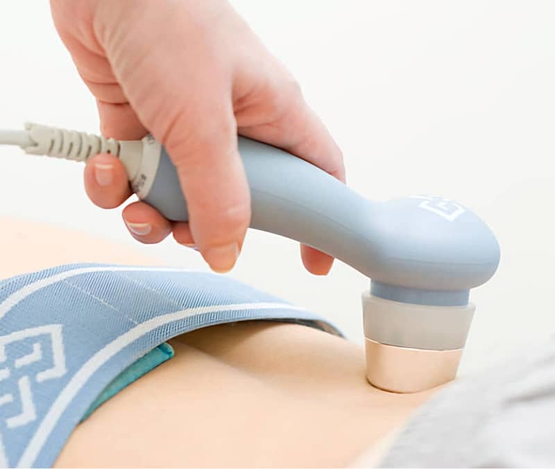 Hand holding a therapeutic ultrasound device applying treatment to a patient's abdomen with sports taping visible, emphasizing injury recovery and pain management at FFC Wellness.