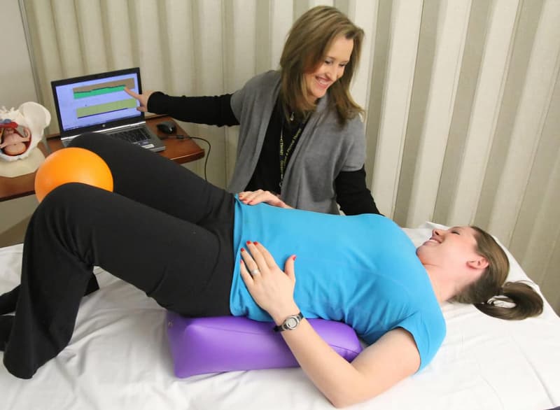 Woman assisting another woman in pelvic floor therapy session using a purple bolster, orange ball, and laptop for biofeedback, highlighting rehabilitation and muscle strengthening.